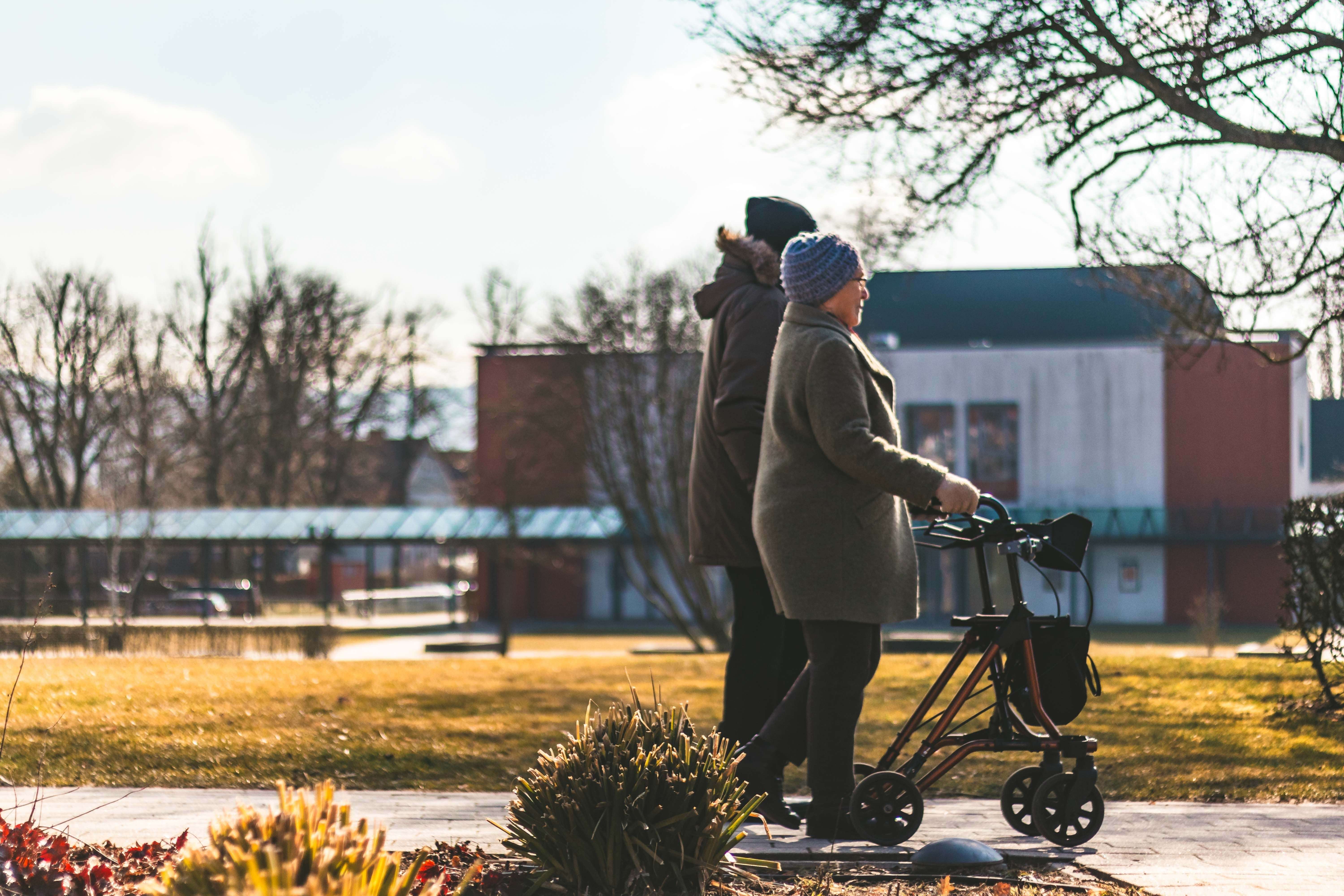 Two senior adults walking with a rollator in a sunny park on a crisp day.