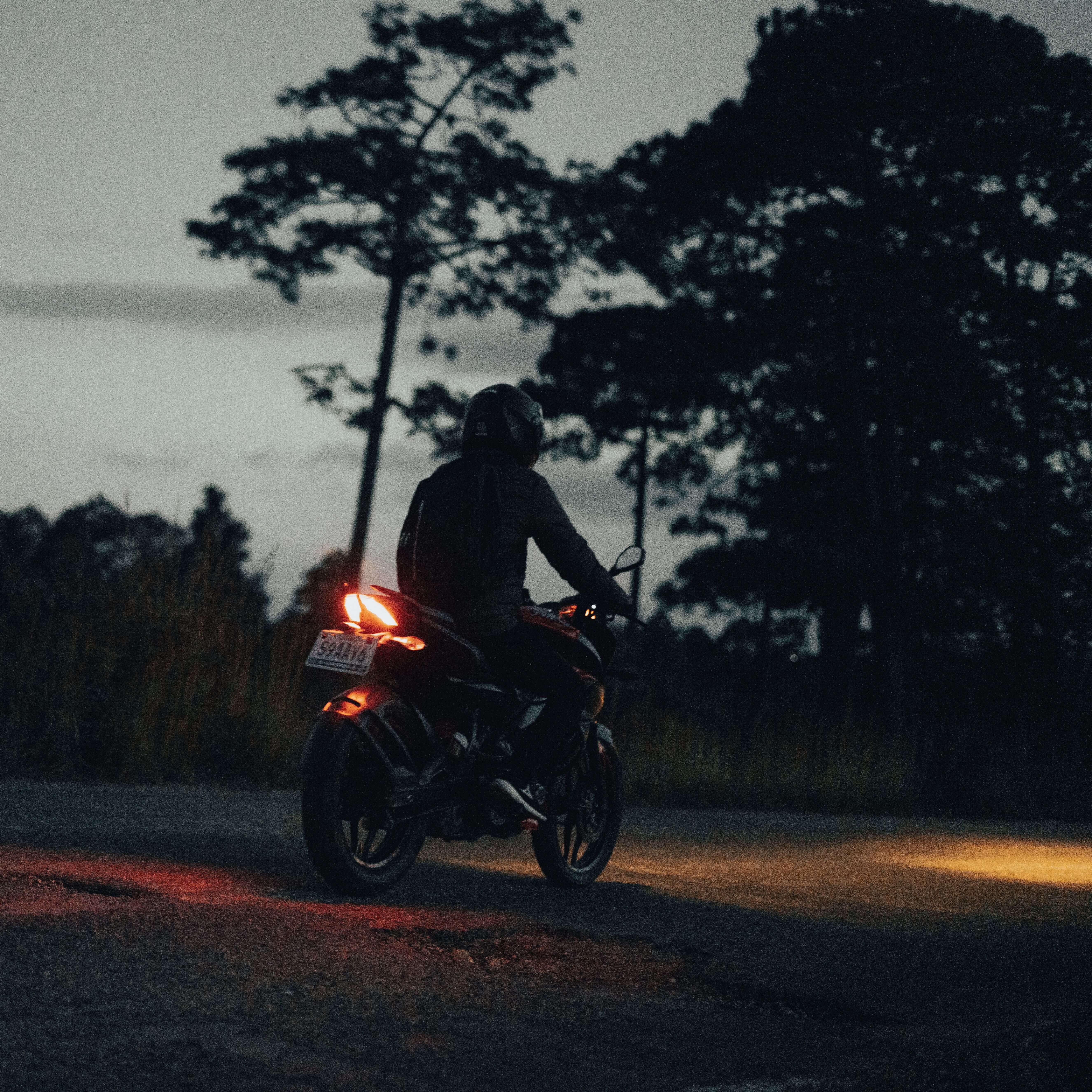 A motorcyclist travels at dusk on a quiet rural road with trees in the background.