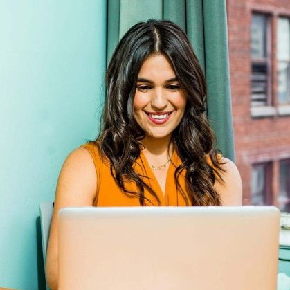 Confident woman in a bright office working on a laptop, surrounded by urban scenery and greenery.