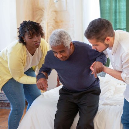 An elderly man receiving assistance from caregivers in a cozy home environment.