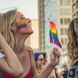 Smiling woman with rainbow face paint waving a flag at a lively Pride parade.