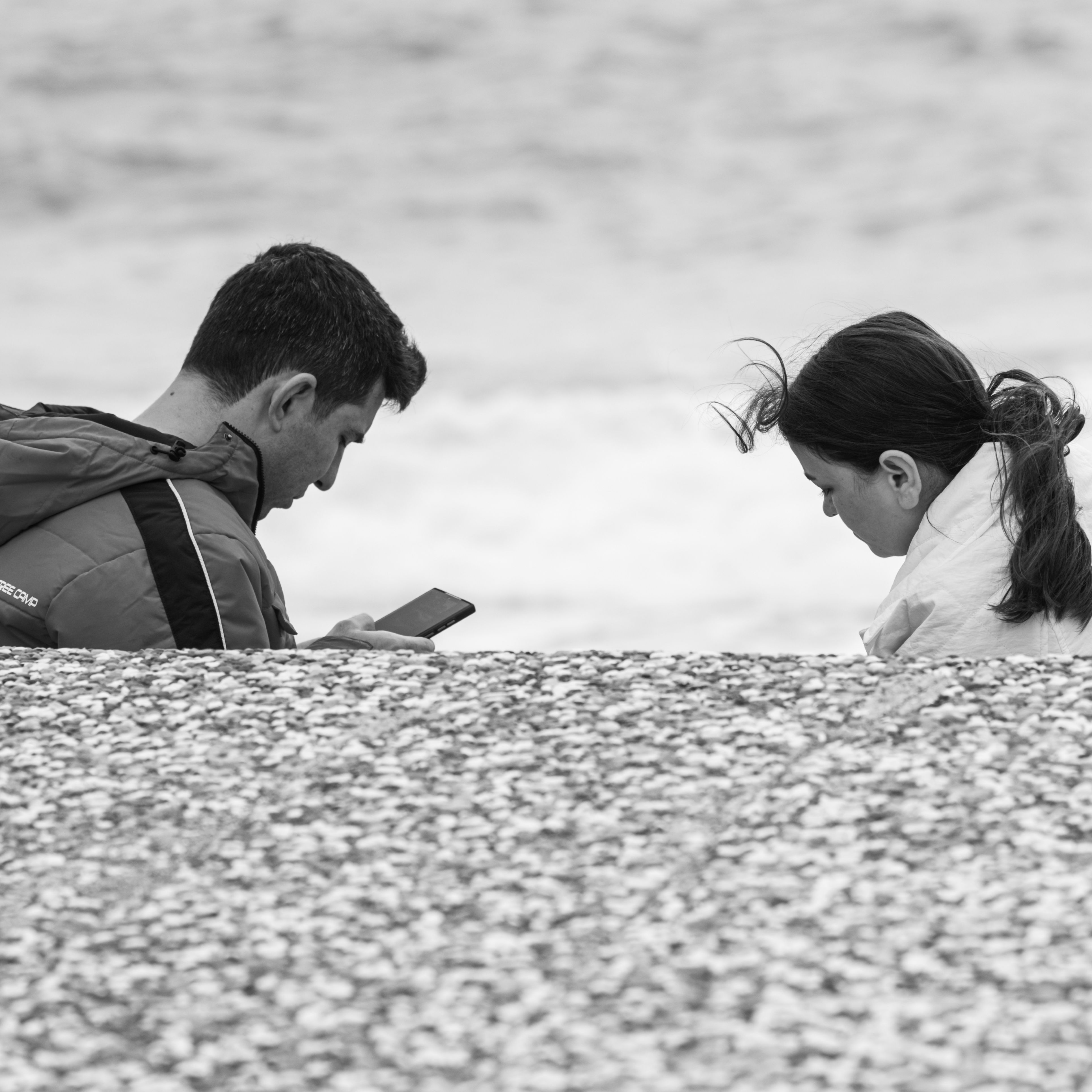 Monochrome image of two adults by the ocean, engaged with a phone, depicting modern connection.