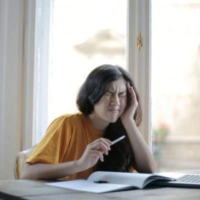 Ethnic female student in casual wear sitting at table with laptop and notebook and grimacing from migraine while doing homework during distance learning