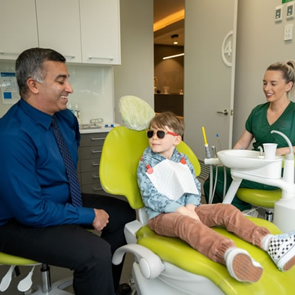 a smiling dentist and his assistant standing on either side of a young patient wearing sunglasses on a dentist chair with dental cabinets in the background