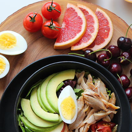 A healthly bowl of salad and other assorted fruits and vegetables around the bowl