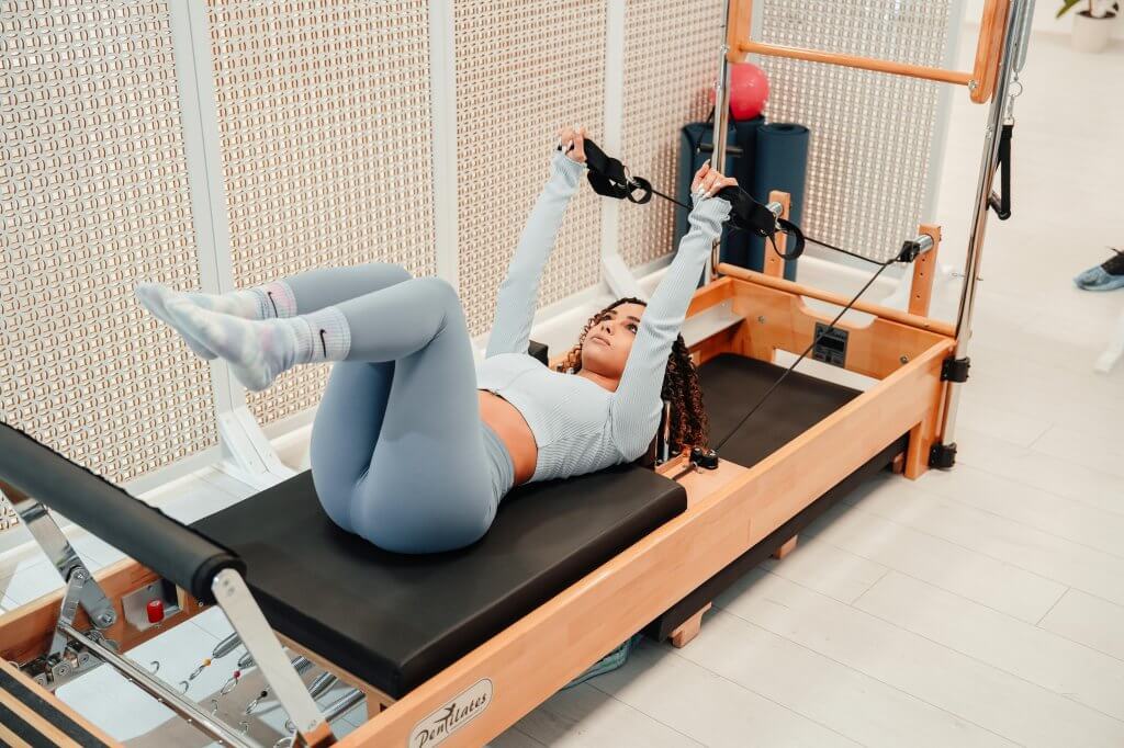 A woman performs pilates exercises on a reformer machine indoors, focusing on fitness and flexibility.