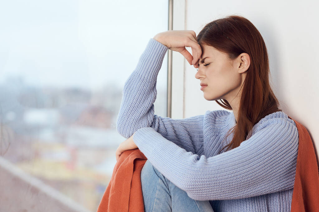 woman with sad expression sits by the window with a plaid