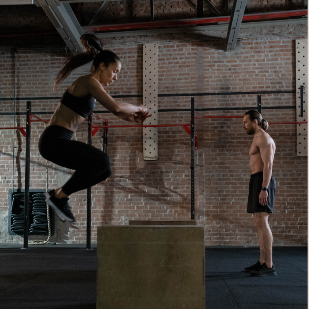 Woman in athletic clothing jumps up onto a large block in a dark, brick exercise space.