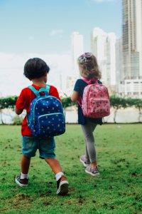 Two kids with colorful backpacks strolling through a city's green park in daylight.