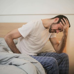 A young man in pajamas holding his head, sitting on a bed, appears to be experiencing a headache.