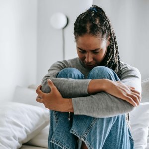 A young woman embraces her knees while sitting on a bed, appearing thoughtful and introspective.