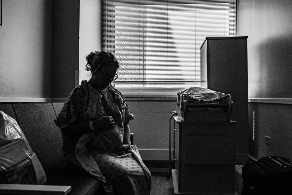 A thoughtful pregnant woman in a hospital room, captured in black and white.