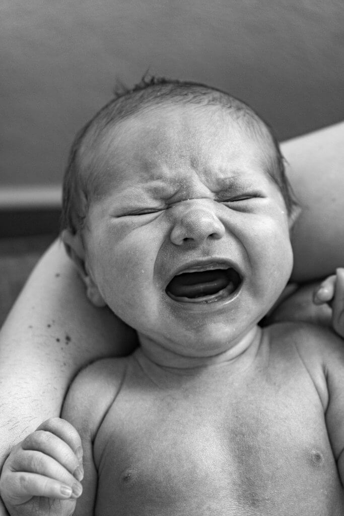 Close-up black and white photograph of a crying newborn being cradled in arms.