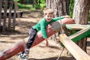 Smiling boy in a green sweater playing on a log in an outdoor park. Fun and leisure in nature.