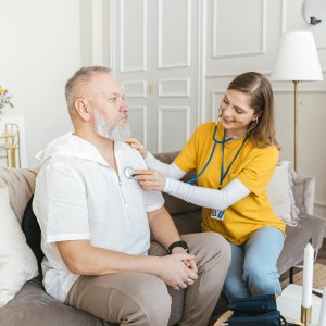 A nurse uses a stethoscope for a home check-up on a senior adult in a cozy living room.