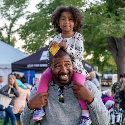 A joyful father carrying his daughter at an outdoor festival surrounded by people and tents.