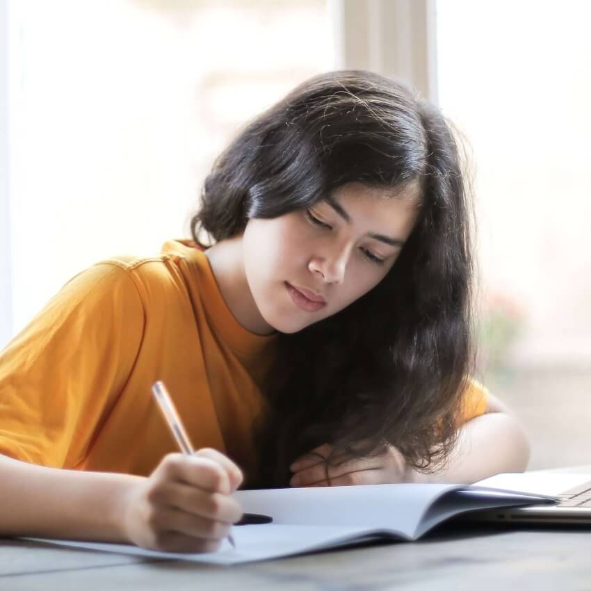 A young woman focused on writing in her notebook near a sunlit window.