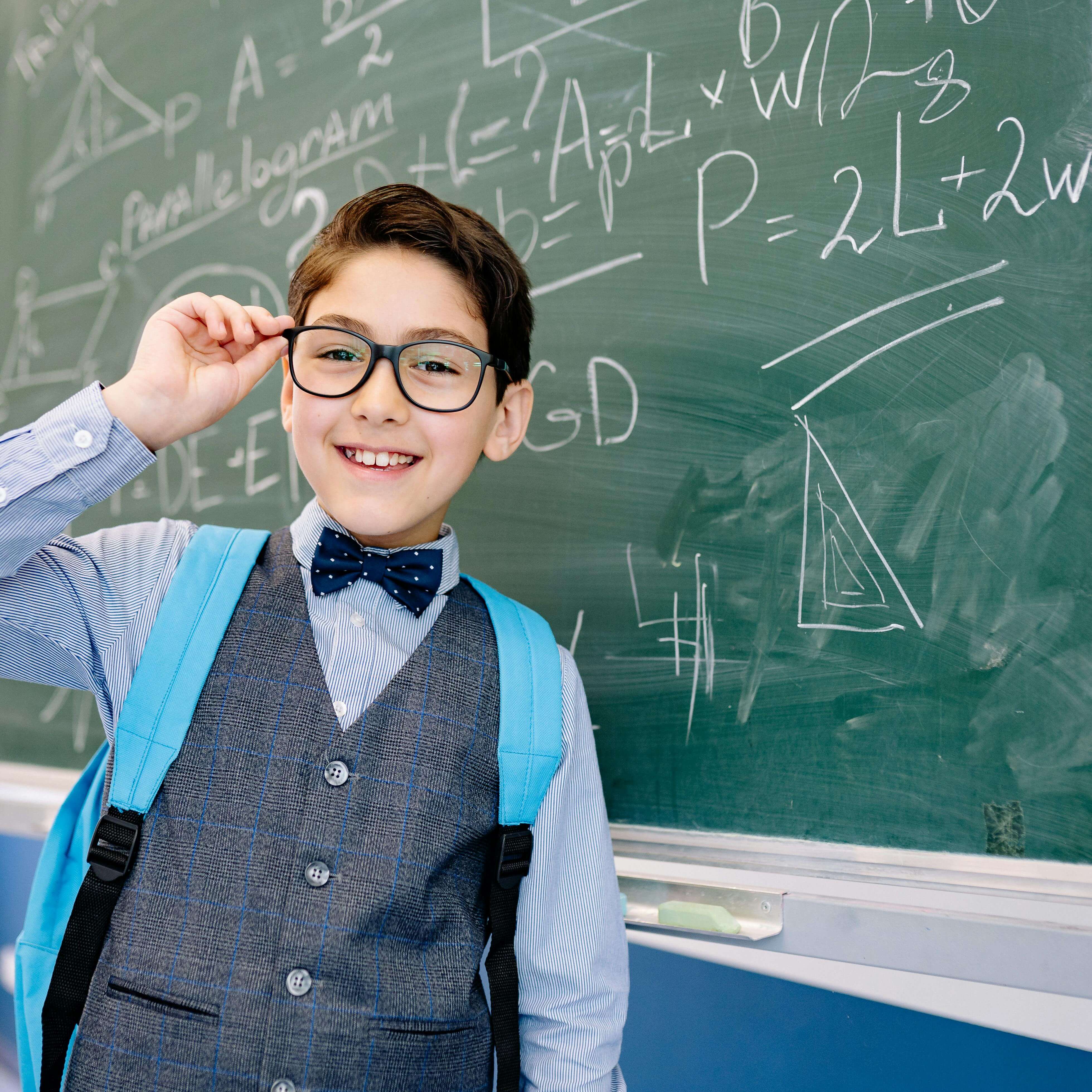 Smiling boy in a bowtie stands by a blackboard with math equations, wearing glasses and a backpack.