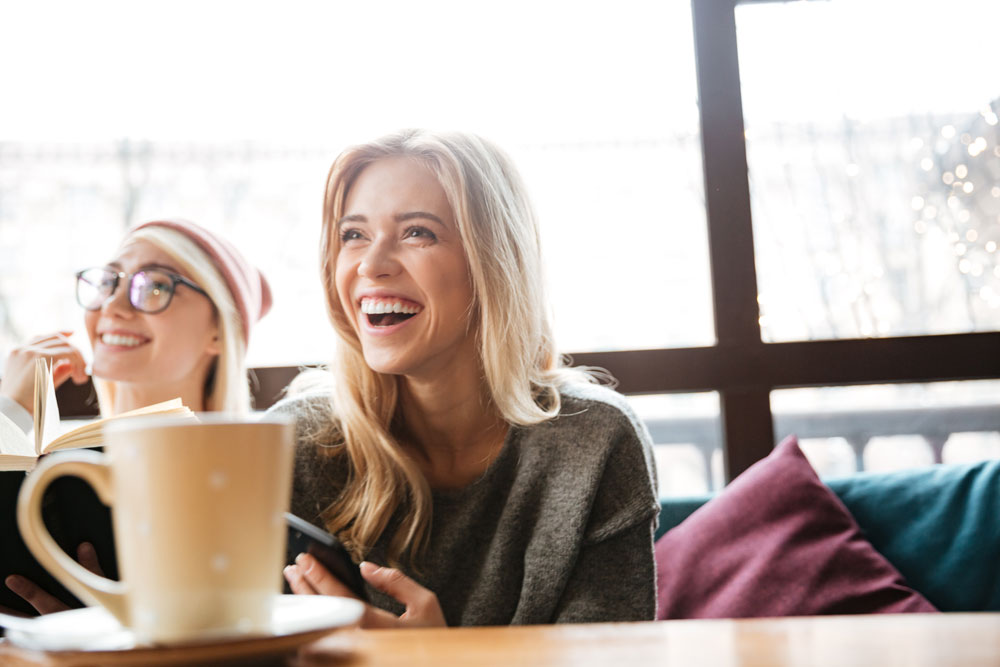 graphicstock-picture-of-young-laughing-two-women-friends-sitting-in-cafe-and-reading-book-while-holding-phone_rJMtvKOAag
