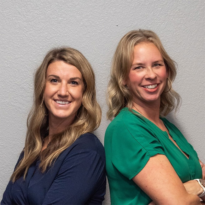 Dr. Lindsay Daniels and Dr. Nicole Des Marais in scrubs standing back to back in front of a white wall at Go Health Chiropractic in Robbinsdale, MN