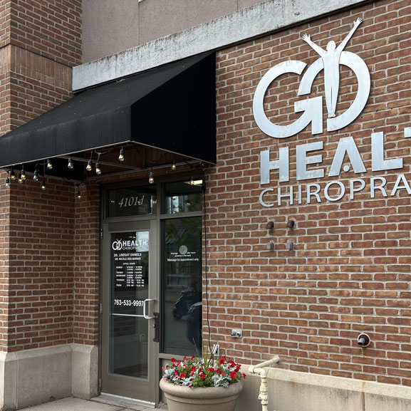 The front door of Go Health Chiropractic with associated hours and the brick building with metallic Go Health Chiropractic lettering in Robbinsdale, MN