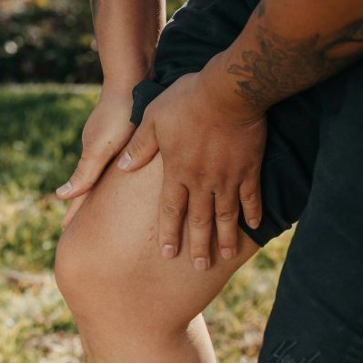 A close-up image of a person holding their knee, indicating pain or injury, with greenery in the background.