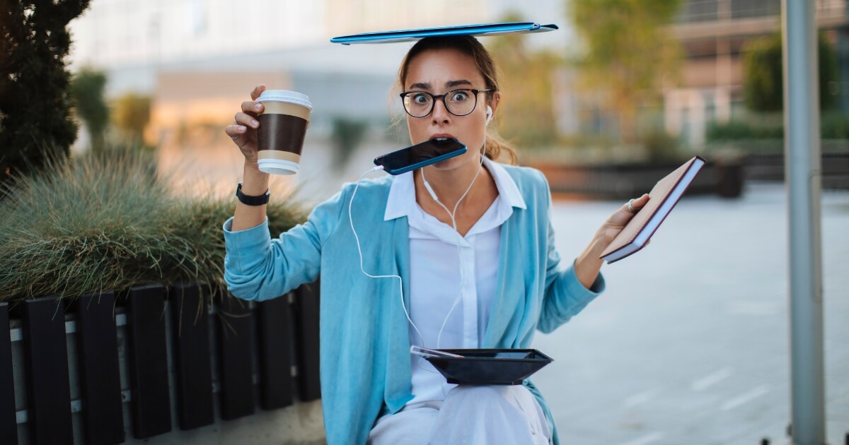 Woman juggling coffee, phone, eating, notes, and laptop.