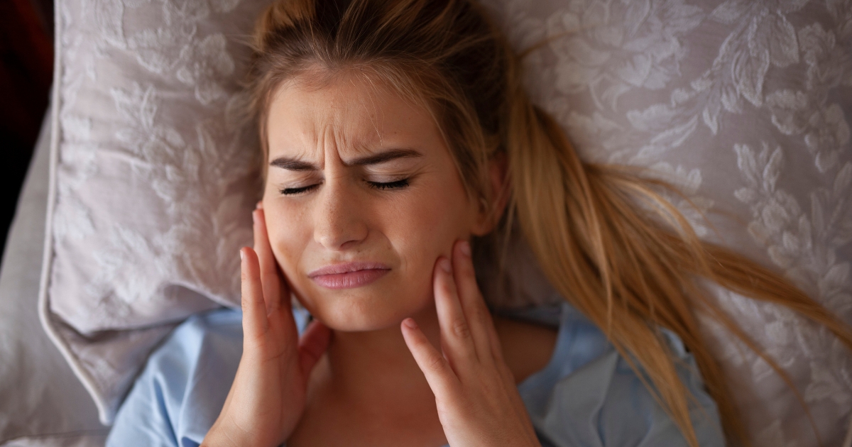 Woman laying in bed and grabbing her jaw in pain.