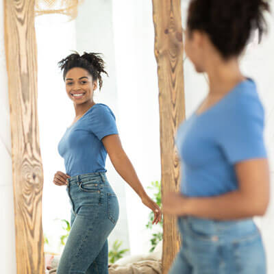 A woman smiling and looking at her waistline in the mirror