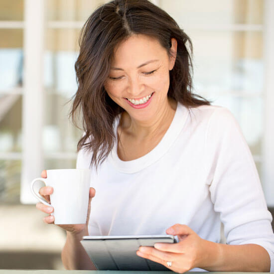 A woman in a white shirt smiles while holding a ceramic coffee cup looks down at an iPad