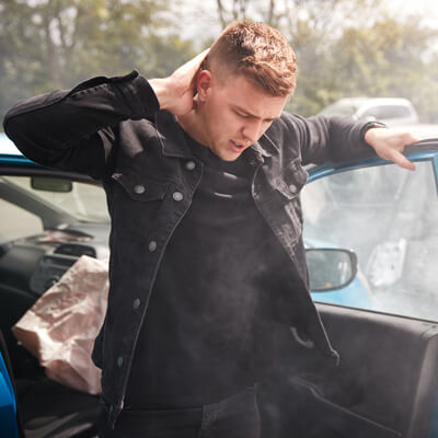 A man standing outside of a smoking car, rubbing his neck.