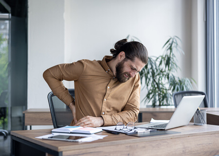 man in office setting suffering from back pain while working at a laptop