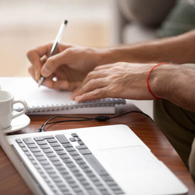 person writing in a notebook at their laptop